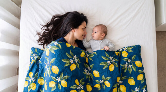 A happy young mother lies on the bed next to her baby under a bright lemon blanket. A touching view from above, filled with family comfort, love and tenderness