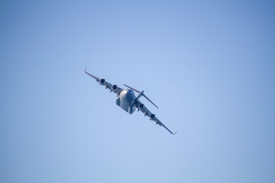 C-17 Globemaster III military transport aircraft in flight, viewed from below against a blue sky with a white cloud below the aircraft, showing its four jet engines and distinctive high-wing design.