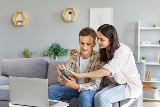 Young family couple using a laptop computer and a smartphone. Man and woman sitting on the couch by a small table with a laptop, looking at their mobile phone, and using an online banking app together