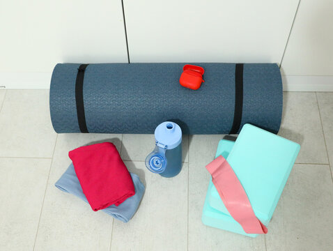 Rolled Blue Yoga Mat With Water Bottle, Resistance Band, Folded Towel, Foam Blocks, And Earbuds Case Arranged On Light Tile Floor For Home Workout, Stretching, Relaxation, And Wellness