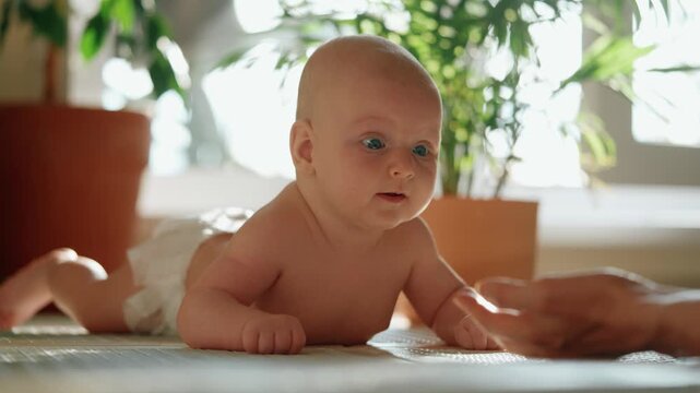 Adorable infant boy with big blue eyes lying on his tummy, practicing head control while his mother gently touches and supports him on a sunlit floor at home