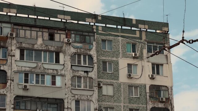 Weathered multi story residential apartment panel building facade with enclosed balconies and overhead trolleybus wires in post soviet Eastern European city under cloudy sky static establishing shot