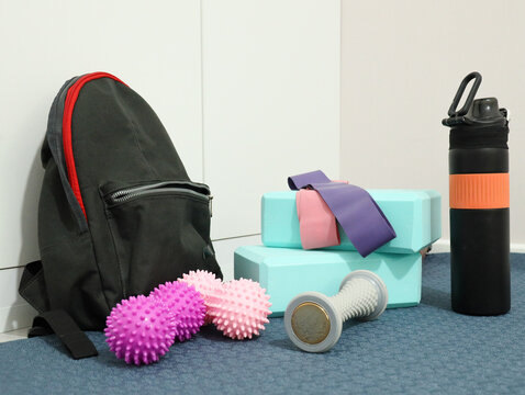 Fitness Equipment Arranged For Home Training And Recovery, Including Resistance Bands, Yoga Blocks, Foam Rollers, Dumbbell, Water Bottle, And Backpack On Blue Mat Beside Clean White Wall