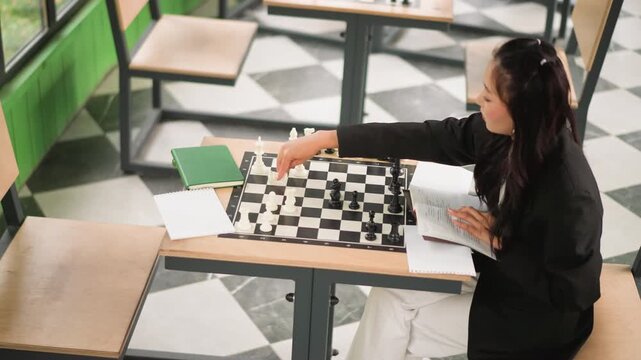 Close up of young woman playing chess at table in classroom setting reading book and moving pieces. Woman wears black jacket with white pants as green notebook rests on desk alongside open