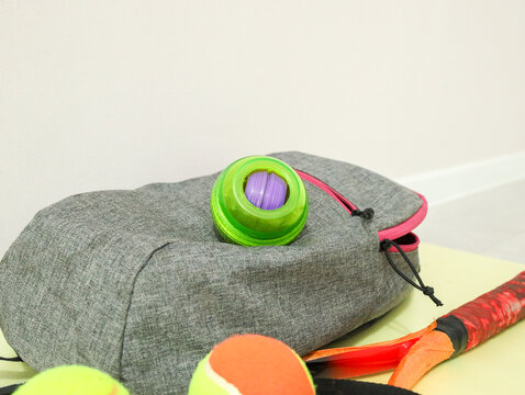 Gray Fabric Backpack With Pink Zipper, Bright Green Toy Container, Colorful Balls, And Sports Gear Resting On Light Surface In Soft Indoor Light, Showing Casual Leisure Equipment Arrangement