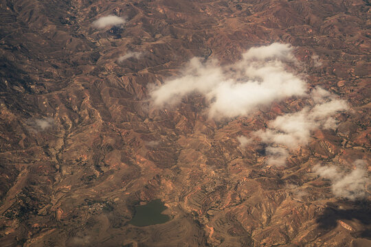Aerial view of the rugged, arid landscape featuring terraced hills, a small lake, and scattered white clouds in Madagascar.
