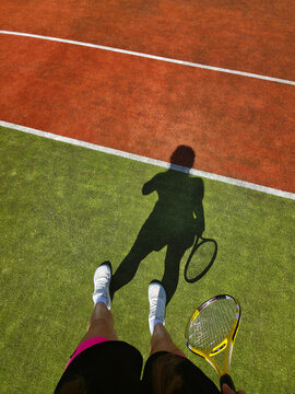 Shadow of a female tennis player holding a racket on a clay court in a vertical format, mobile photography style, athletic woman standing on lines, top view of legs in sneakers, creative sports