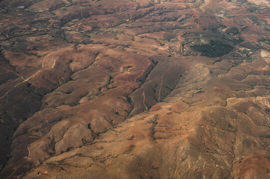 Aerial view of the arid, hilly landscape of Madagascar, featuring reddish-brown soil, winding dirt paths, and sparse vegetation seen during a flight from Morondava to Antananarivo.