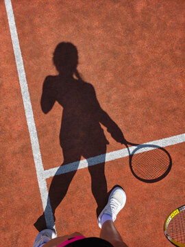 Shadow of a female tennis player holding a racket on a clay court in a vertical format, mobile photography style, athletic woman standing on lines, top view of legs in sneakers, creative sports