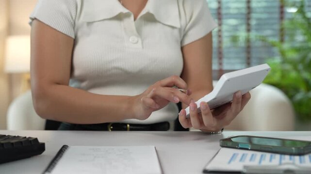 Young businesswoman managing finances at her desk, calculating expenses with a calculator, and writing notes in a notebook, focused on budgeting, accounting, and financial planning