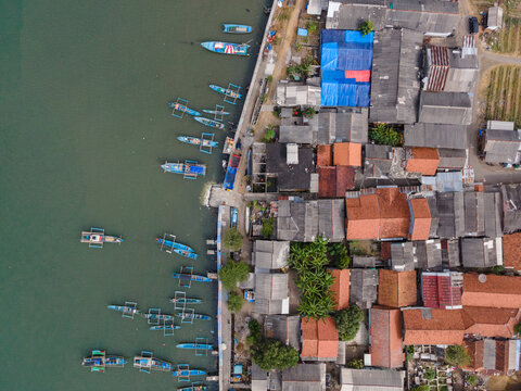 Aerial view of fishing boats moored along the coastline next to a dense residential area with terracotta rooftops in Cilacap, Central Java, Indonesia.
