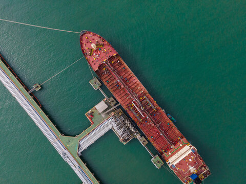 Aerial view of a large oil tanker ship docked at an industrial terminal with complex piping systems and mooring lines in the green waters of Cilacap, Central Java, Indonesia.