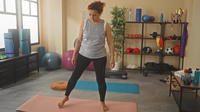 Middle aged woman stretching on yoga mat in studio with side bend, hand to thigh and bare feet grounded; serenity mindfulness wellbeing.