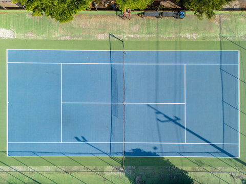 Aerial view of a blue tennis court with white lines and a net, surrounded by green grounds with shadows cast by poles in Klaten, Central Java, Indonesia.