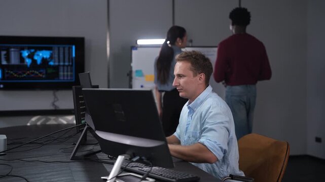 Caucasian male programmer coding on computer monitors in modern IT office, Software developer working on future technology business innovation while diverse colleagues brainstorming in background