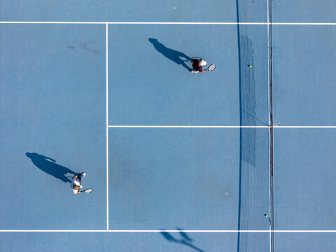 Aerial view of two tennis players on a blue hard court with long shadows cast on the surface in Klaten, Central Java, Indonesia.