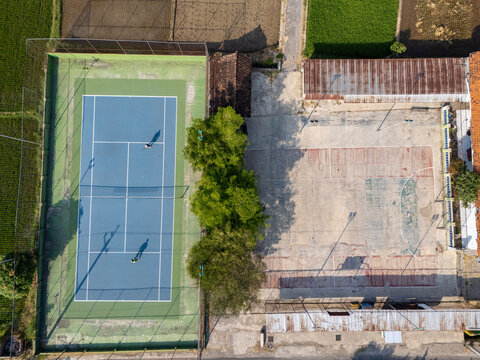 Aerial view of a blue tennis court and an adjacent concrete sports court surrounded by lush trees and agricultural fields in Klaten, Central Java, Indonesia.