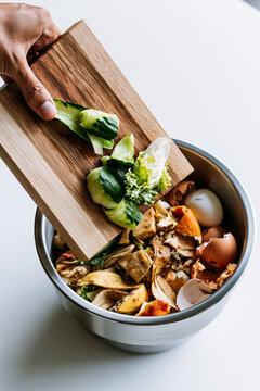 Top-down shot of a person's hand holding a cutting board over a stainless steel bowl filled with organic kitchen food scraps for composting.
