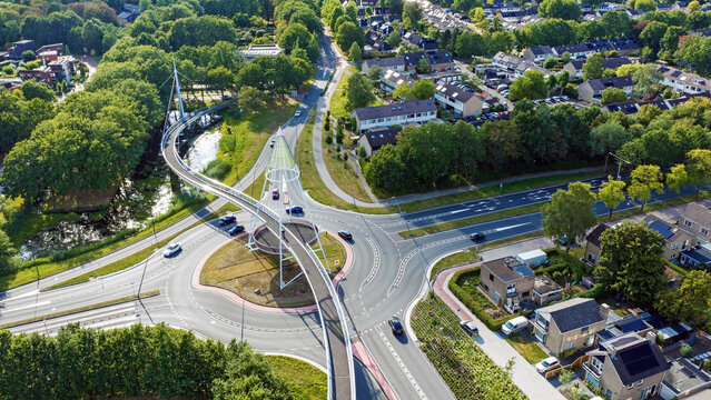 Aerial view of De Slinger Bicycle bridge spanning over a roundabout in a residential area with lush green trees and suburban houses in Drachten, Friesland, Nederland.