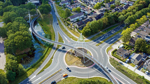 Aerial view of De Slinger Bicycle bridge spanning over a modern roundabout with surrounding residential houses and lush green trees in Drachten, Friesland, Netherlands.