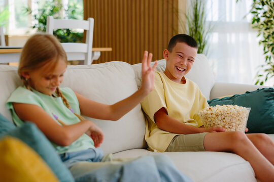 Brother and sister sharing popcorn and laughter on a cozy living room couch, enjoying a bright, playful family moment watching a movie and bonding together