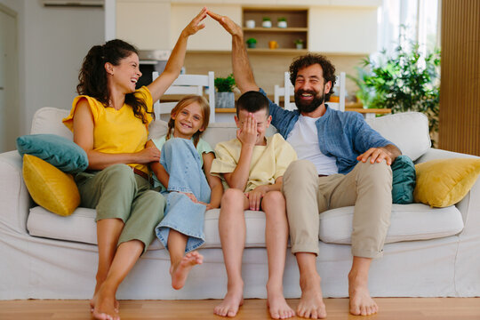 Young family on a cozy living room couch smiling and laughing as parents form a house shape with their hands, symbolizing home, protection, security and togetherness
