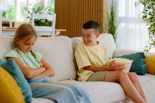 Young brother laughing while holding a bowl of popcorn and teasing his upset sister with crossed arms, creating a classic sibling rivalry scene at home on the sofa