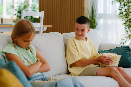 Two siblings on a sofa in the living room during a petty argument: boy teasing while sister clutches a bowl of popcorn, upset and sulking in a real-life family moment