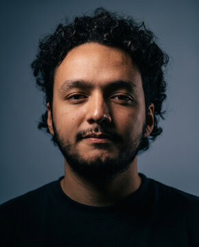 Close up portrait of a young man with curly hair and a beard looking directly at the camera.