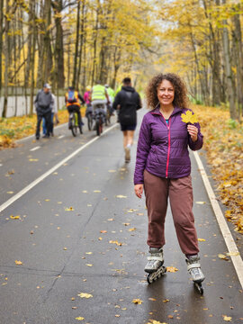 Woman rollerblades autumn park path. Fall leaves scatter asphalt surface. Group cyclists ride behind her. Trees frame the autumnal scene. Person smiles, holds a leaf