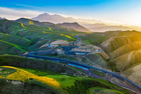 Winding highway traverses rolling green hills and rugged terrain beneath majestic snow peaks in Xinjiang, China.