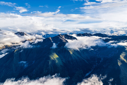 Majestic mountain range emerging through rolling white clouds under a bright blue sky, showcasing dramatic alpine peaks and deep valleys.