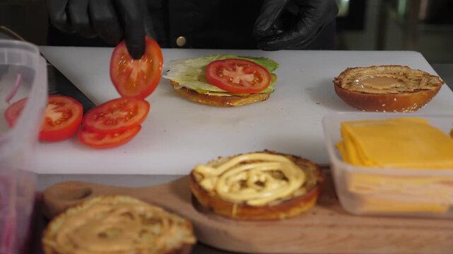 The chef is preparing a burger, he places a slice of tomato on a bun with sauce. The process of making burgers takes place in the kitchen of the restaurant, and there are many ingredients on the table