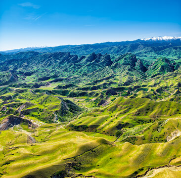 Lush green rolling hills and dramatic geological rock formations stretching toward snow-capped mountains under a clear blue sky.