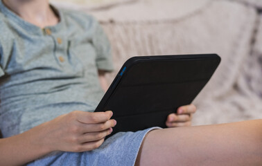 Young boy holding digital tablet sitting on couch at home. Child studying online, playing games or watching video content on modern portable device indoors. Technology and education concept. ( © Тетяна Хіміченко