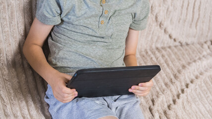 Young boy holding digital tablet sitting on couch at home. Child studying online, playing games or watching video content on modern portable device indoors. Technology and education concept. ( © Тетяна Хіміченко