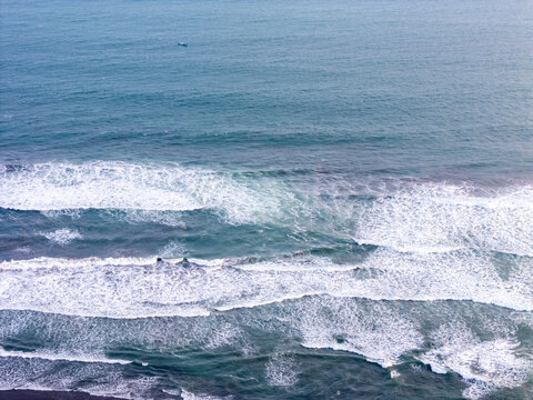 Aerial view of ocean waves crashing onto a dark sand beach with a small boat floating in the distance in Bantul, Special Region of Yogyakarta, Indonesia.
