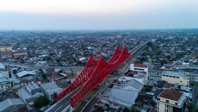 Aerial view of a prominent red truss bridge spanning over a road in a dense urban neighborhood in Banjarsari, Central Java, Indonesia.