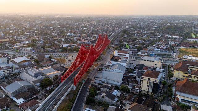 Aerial view of the red steel arch Tirtonadi Bridge spanning over the urban landscape with dense rooftops and railway tracks in the golden hour light in Banjarsari, Central Java, Indonesia.