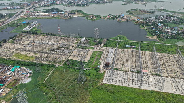 Aerial view of a power substation with electrical transformers and high voltage transmission towers situated near a lake and green landscape in Dhaka, Dhaka Division, Bangladesh.