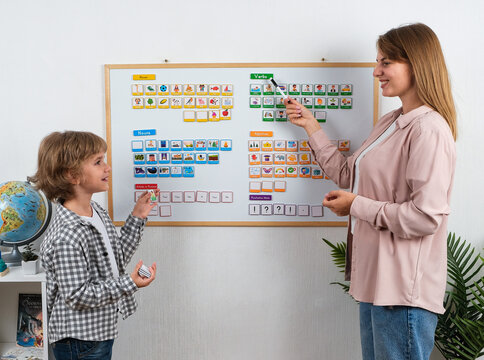 Female teacher points to English grammar cards on a magnetic learning board while teaching a schoolboy parts of speech, vocabulary and sentence building in a classroom.