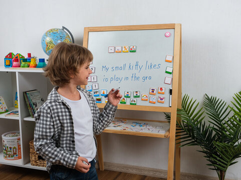 Schoolboy writes an English sentence on a learning board while using colorful word and picture cards as visual aids for practicing grammar, vocabulary and sentence building.