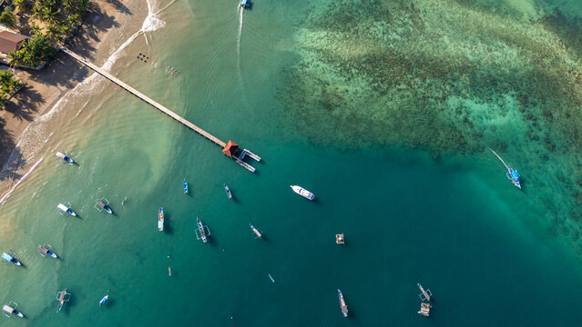 Aerial view of Pantai Karanggongso featuring a long wooden pier extending into the turquoise bay, surrounded by numerous small fishing boats and sandy coastline in Watulimo, East Java, Indonesia.
