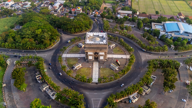 Aerial view of Simpang Lima Gumul monument situated in a large roundabout surrounded by lush trees and urban roads in Kediri, East Java, Indonesia.