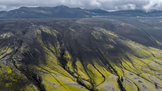 Aerial view of the volcanic landscape of Raudaskal crater with dark lava fields and vibrant green moss under a cloudy sky in Hella, Rangarthing ytra, Iceland.