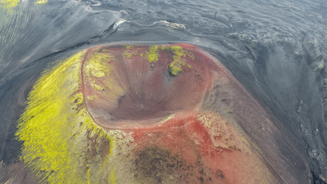 Aerial view of Raudaskal crater with its distinctive red volcanic soil and vibrant green moss, set against a vast landscape of dark basaltic lava fields in Hella, Rangarthing ytra, Iceland.