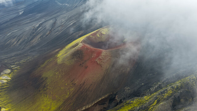 Aerial view of the vibrant red Raudaskal crater surrounded by mossy green slopes and dark volcanic ash, partially obscured by low-hanging clouds in Hella, Rangarthing ytra, Iceland.