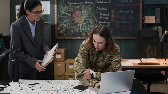 Young adult Caucasian female soldier in camouflage typing on laptop with middle-aged Caucasian female colleague in military office. Ideal for defense industry marketing or security training