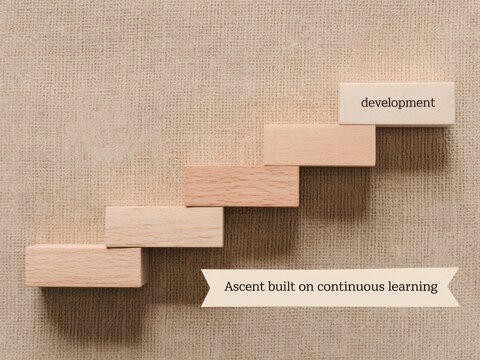 Wooden blocks forming a rising staircase on textured beige fabric, with text development and a ribbon stating Ascent built on continuous learning. Factual flat lay composition.