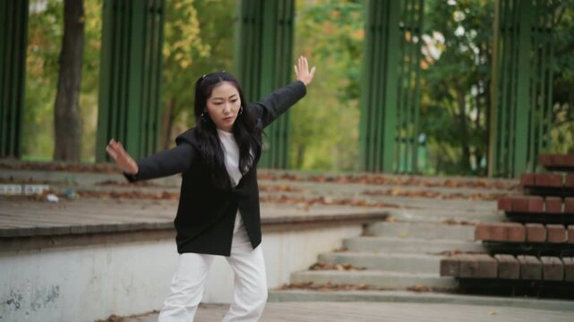 Close up of young woman performing tai chi movements in outdoor park setting. Woman wears black blazer and white pants gracefully moving through poses with fluid arm gestures against green